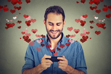 Portrait happy man sending love sms text message on mobile phone with red hearts flying away from screen isolated on grey wall background. Human emotions