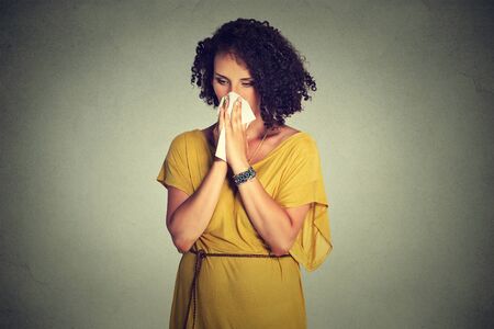Middle aged woman sneezing in a tissue blowing her runny nose standing isolated on gray wall backgroundの写真素材