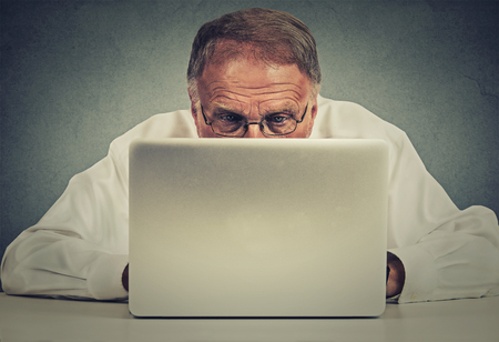 Closeup portrait elderly man sitting at table working on laptop computer isolated on gray office...