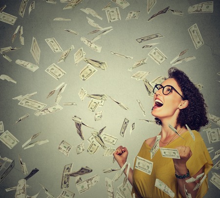 Portrait happy woman in glasses exults pumping fists ecstatic celebrates success under a money rain falling down dollar bills banknotes isolated on gray wall background with copy space Portrait happy woman in glasses exults pumping fists ecstatic celebrates success under a money rain falling down dollar bills banknotes isolated on gray wall background with copy space