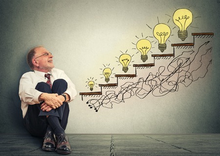 Excited happy elderly executive man relaxed sitting on a floor in his office looking up at business success company growth graph made of light bulbs isolated on gray wall texture backgroundの写真素材