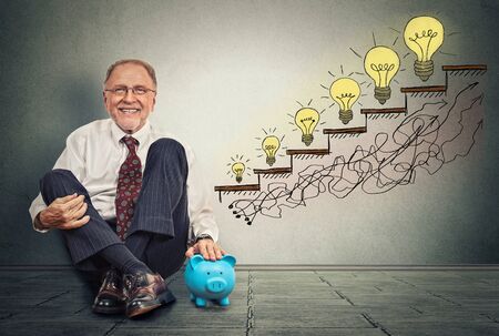 Excited happy senior executive man sitting on a floor in his office with piggy bank celebrates business success, promotion, company growth isolated on gray wall texture backgroundの写真素材