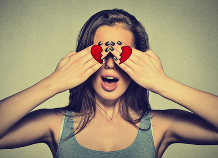 Beautiful young woman eyes covered by hands with broken heart printed on them. Girl in love isolated on gray wall backgroundの写真素材