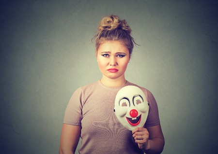 Portrait young upset worried woman with sad expression holding a clown mask expressing cheerfulness happiness isolated on gray wall background. Human emotionsの写真素材