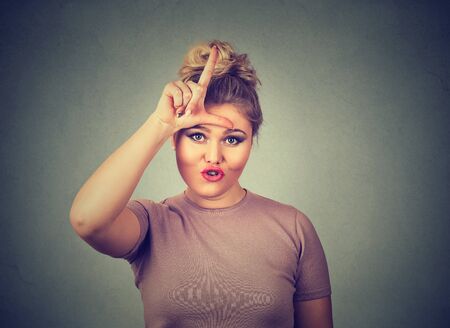 Closeup portrait young unhappy woman giving loser sign on forehead, looking at you, disgust on face isolated on gray wall background. Negative human emotion facial expressionの写真素材