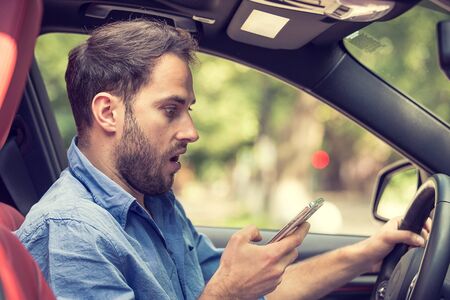 Man sitting in car with mobile phone in hand texting while driving. Distracted shocked guy checking...
