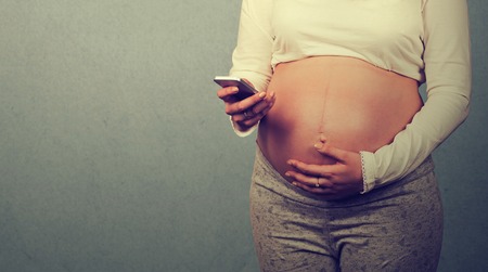 Cropped closeup image young pregnant woman texting on mobile phone isolated on gray wall background...