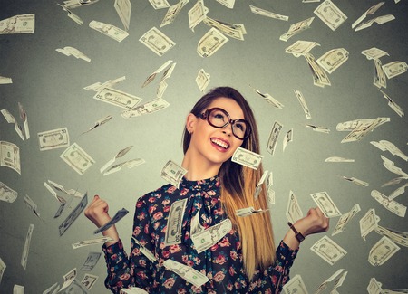 Portrait happy young woman exults pumping fists ecstatic celebrates success under a money rain falling down dollar bills banknotes isolated on gray wall background with copy spaceの写真素材