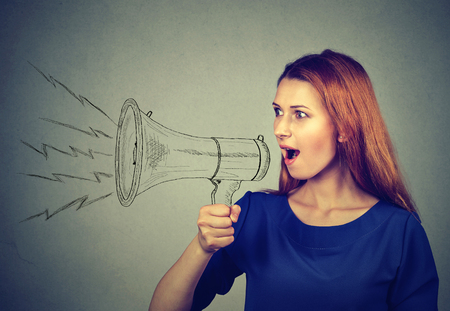 Portrait angry young woman screaming in megaphone isolated on grey wall background. Negative face expression emotion feelings. Propaganda, breaking news, power, social media communication conceptの写真素材