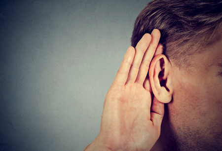 man holds his hand near ear and listens carefully isolated on gray wall background