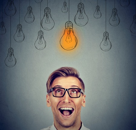 Excited happy man in glasses looking up with light idea bulb sign above head has an idea isolated on gray wall backgroundの写真素材