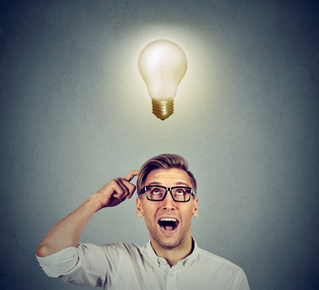 Happy man in glasses looking up at bright light idea bulb above head isolated on gray wall backgroundの写真素材