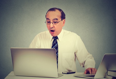 Middle aged shocked business man sitting in front of laptop computer looking at screen isolated grey background. Human face expression emotion feelings problem perception reactionの写真素材