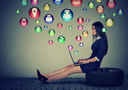 Young woman sitting on a tire working on laptop computer with social media application icons flying out of screen. Modern technology and people conceptの写真素材