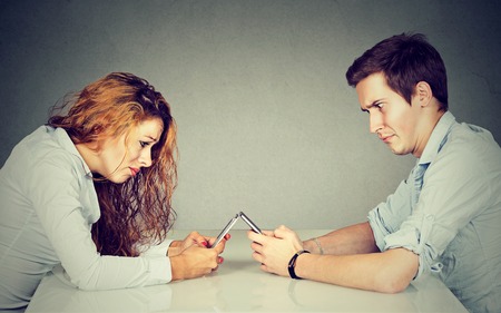 Smartphone addiction concept. Frustrated annoyed young woman and man sitting at table with smartphone and do not looking at each other ignoringの写真素材