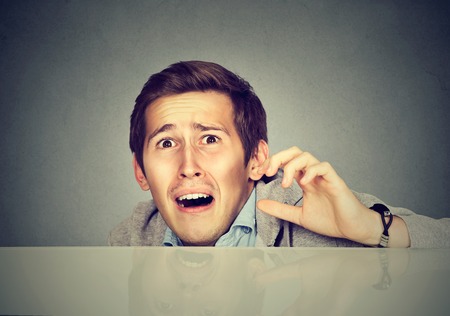 Funny scared man peeking from behind the desk isolated on gray backgroundの写真素材