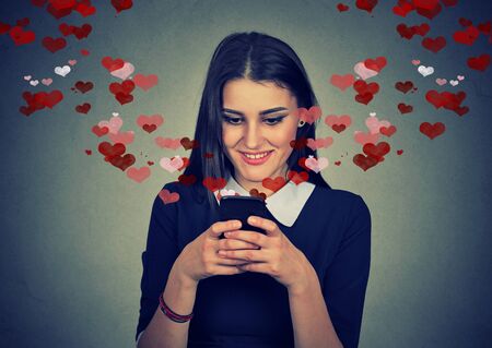 Portrait happy woman sending love sms text message on mobile phone with red hearts flying away from screen isolated on gray wall background. Human emotions feelingsの写真素材