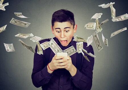 Technology online banking money transfer, e-commerce concept. Amazed young man using smartphone with dollar bills flying away from screen isolated on gray wall background.の写真素材