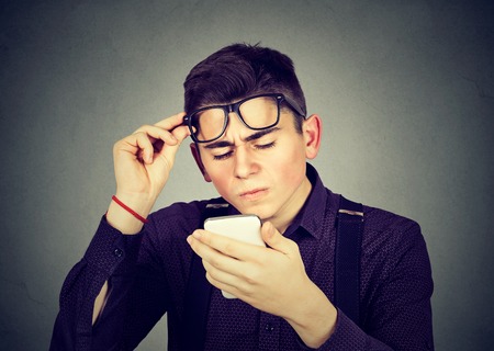 Young man with glasses having trouble seeing cell phone has vision problems. Bad text message. Negative human emotion facial expression perceptionの写真素材