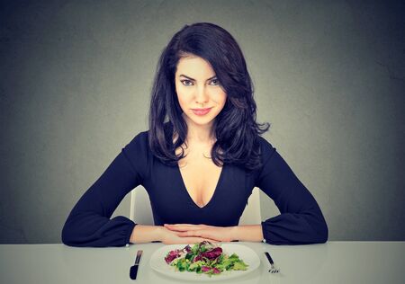 Attractive young woman sitting at table ready to eat green salad isolated on gray backgroundの写真素材