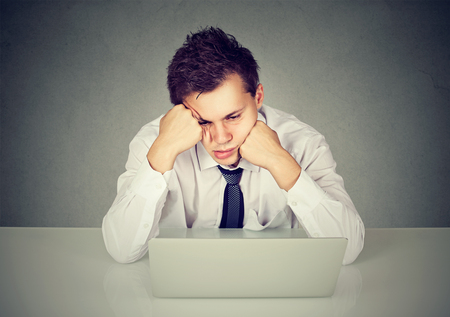 Overworked young bored man sitting at desk in front of his laptop computer looking down の写真素材