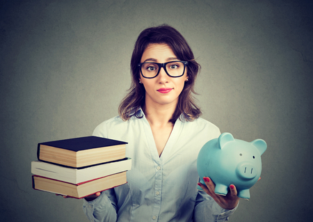 Student loan concept. Young woman with stack pile of books and piggy bank full of debt rethinking future career path Student loan concept. Young woman with stack pile of books and piggy bank full of debt rethinking future career path