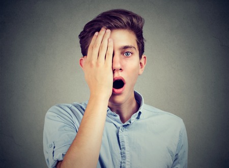 Surprised man, with one eye, covered by hand looking at camera isolated on gray background