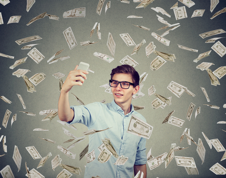 Young man in eyeglasses taking selfie in flying dollar making money by being popular in social...