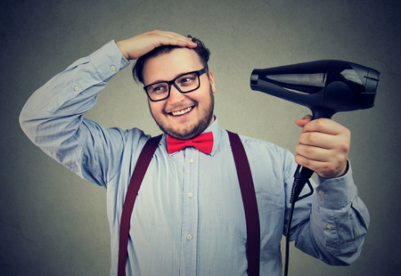 Young chunky man in formal outfit using hairdryer to style hair while posing on gray. の写真素材