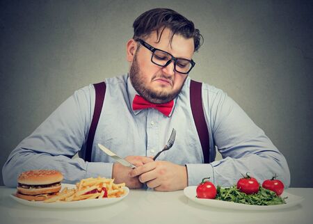 Overweight man in formal outfit looking at saladâ with hate while craving juice hamburger.の写真素材