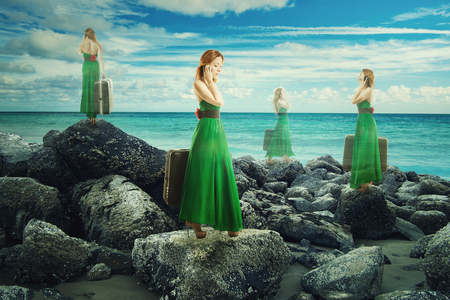 Translucent woman in green dress with suitcase walking in infinite time on rocky shoreline. の写真素材