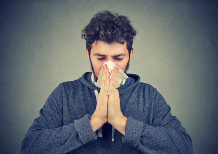 Young man having running nose and blowing it on handkerchief suffering from flu. の写真素材