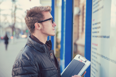 Side view of handsome student in glasses holding document and pad looking on information board outside. の写真素材