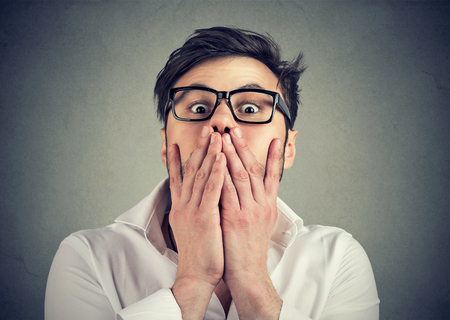 Young man in eyeglasses looking extremely frightened and covering mouth while looking at camera. の写真素材