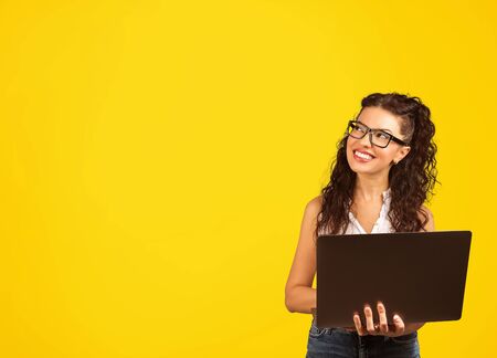 Young positive cool lady with curly hair holding laptop looding away daydreaming and smiling isolated over yellow background の写真素材