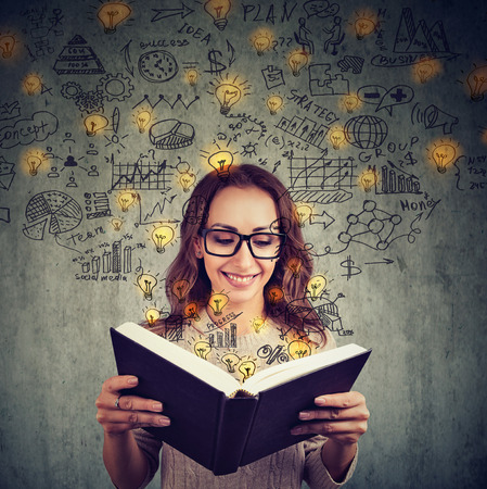 Young girl in glasses reading big book and having plenty of successful ideas looking happy on gray...