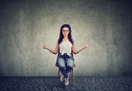 Casual woman in glasses sitting on chair and holding hands apart in meditation and harmony keeping eyes closed. の写真素材