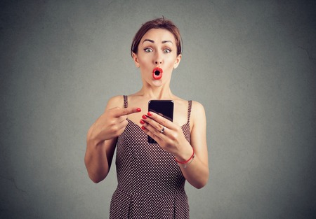 Shocked ironic girl holding smartphone and pointing at it sunned with breaking newsの写真素材