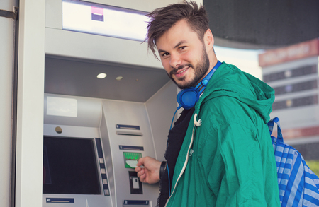 Young hipster guy in headphones smiling at camera while inserting credit card in ATM machine getting cash moneyの写真素材