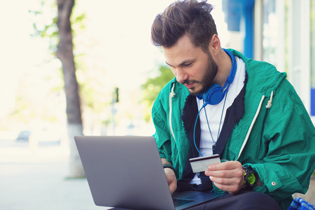Stylish hipster man sitting on street with laptop on knees and doing online purchase with credit...