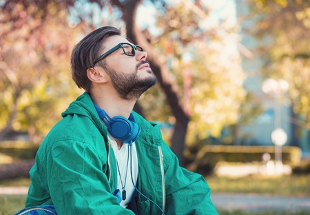 Hipster man in headphones and glasses keeping eyes closed and enjoying fresh air in summer parkの写真素材