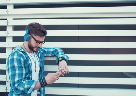 Young stylish man in headphones standing outside and using smartphone while listening to musicの写真素材