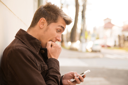 Side view of nervous scared man biting fist while looking at smartphone and standing on streetの写真素材