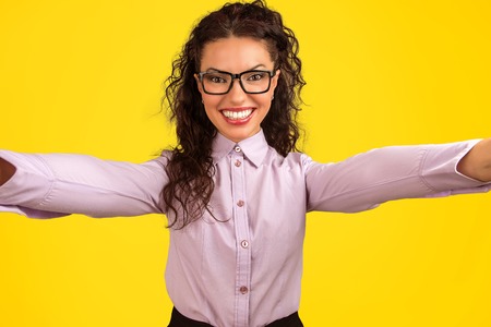 Cheerful young woman in glasses looking at camera and taking selfie on bright yellow background.の写真素材