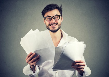 Happy young man in glasses looking through paper documents with good results and smiling on gray backgroundの写真素材