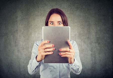 Young woman holding tablet in front of face looking amazed and shocked with news standing on gray...