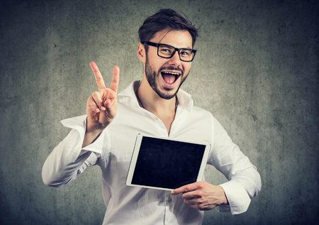 Cheerful model man in white shirt holding new tablet and showing two fingers while looking at camera on gray backgroundの写真素材