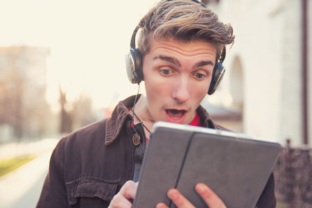 Modern young man in headphones surfing tablet in amazement standing on streetの写真素材