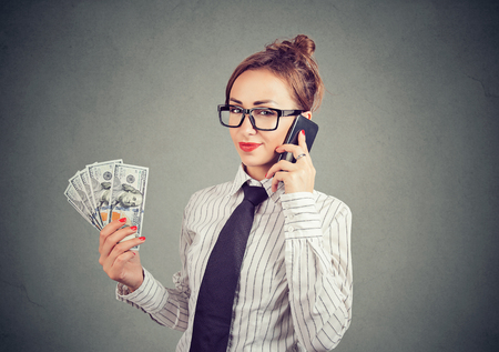 Young satisfied woman in glasses holding dollar bills and speaking on smartphone looking at camera on gray backgroundの写真素材