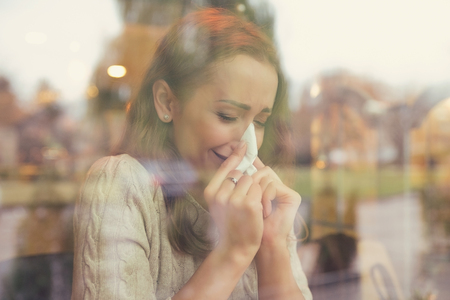 Woman with cold symptoms blowing her nose sneezing sitting in a coffee shop on a rainy autumn dayの写真素材